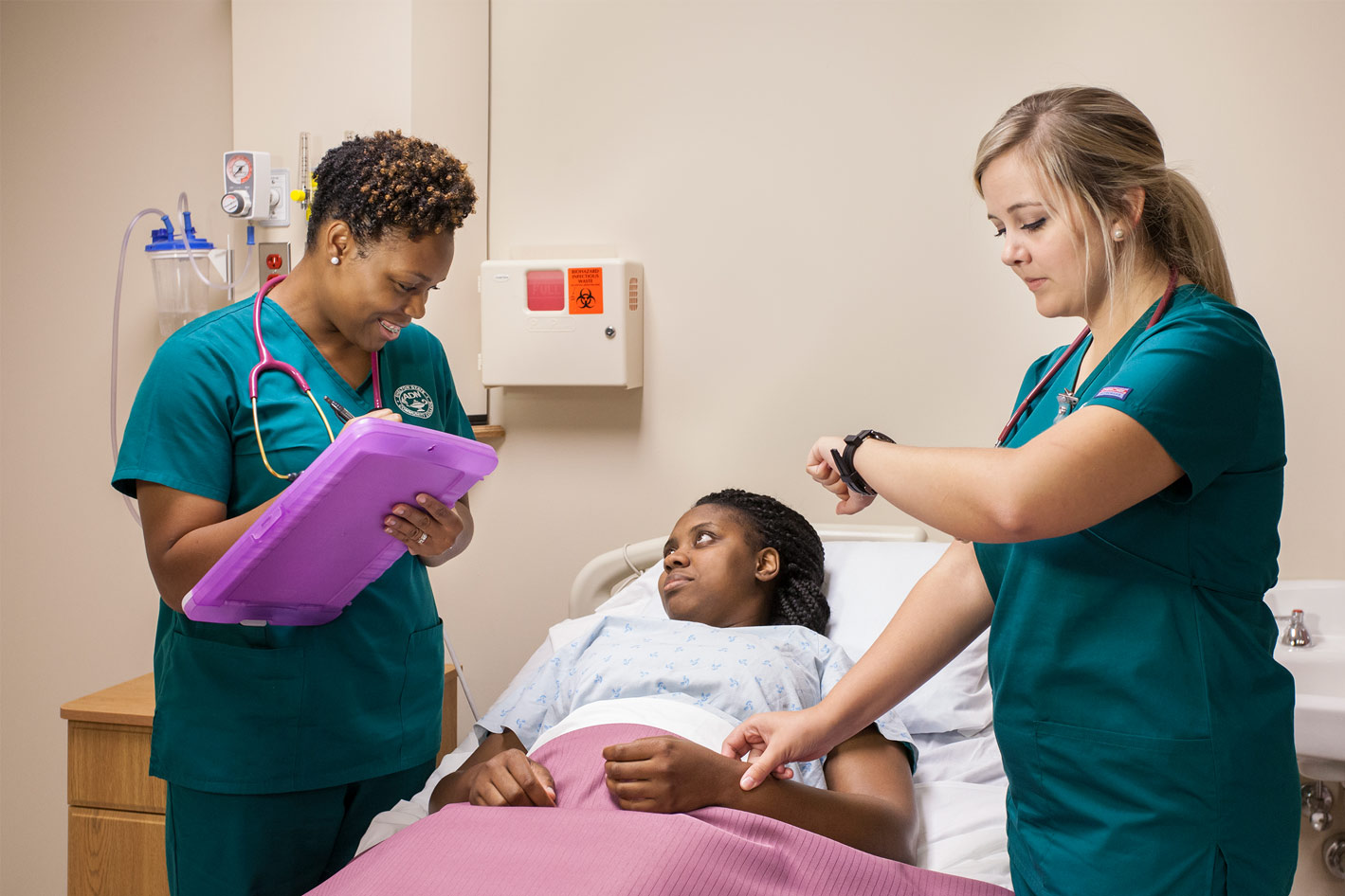 Shelton State Nurses checking a patient. One is checking the patients pulse while the other writes on clipboard